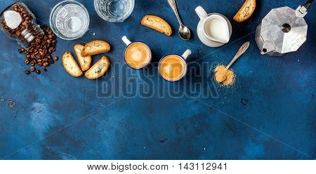 Coffee espresso in cups with italian cantucci, cookies and milk in jug over dark blue painted plywood background, top view, copy space. Food frame concept