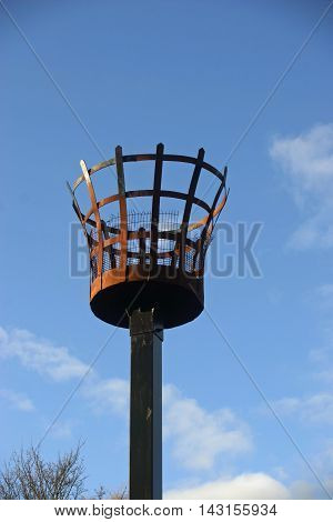Beacon to be used with a fire. Outer metal basket construction with an inner basket for the fire. Metalwork rusted. On a wooden post. Background of blue sky with white cloud and tree without leaves in the lower left corner.