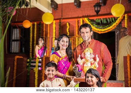 Smart Indian couple holding Ganesh idol or ganpati idol at entrance, welcoming lord ganesh on ganesh festival or chaturthi with kids and parents, happiness concept