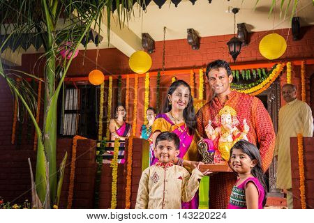 Smart Indian couple holding Ganesh idol or ganpati idol at entrance, welcoming lord ganesh on ganesh festival or chaturthi with kids and parents, happiness concept