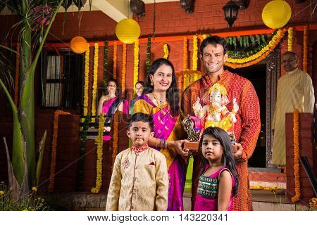 Smart Indian couple holding Ganesh idol or ganpati idol at entrance, welcoming lord ganesh on ganesh festival or chaturthi with kids and parents, happiness concept