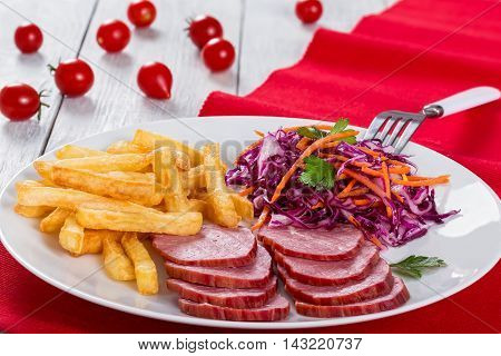 sliced smoked veal fillet french fries and red cabbage salad with carrots cut into strips and parsley dressing with vinegar and olive oil on table mat with cherry tomatoes on white rustic boards close-up