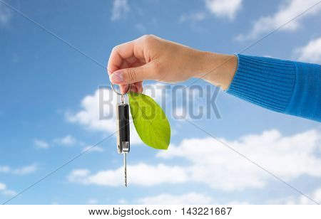 conservation, environment, people, transport and ecology concept - close up of hand holding car key with green leaf trinket over blue sky and clouds background