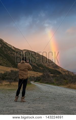 Man taking photo of rainbow over mountain near Lake Pearson / Moana Rua Wildlife Refuge located in Craigieburn Forest Park in Canterbury region South Island of New Zealand