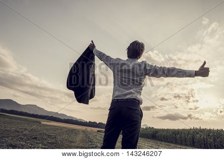 Rear view of single businessman with removed jacket with his arms outspread showing thumbs up sign at the open field during sunset time.