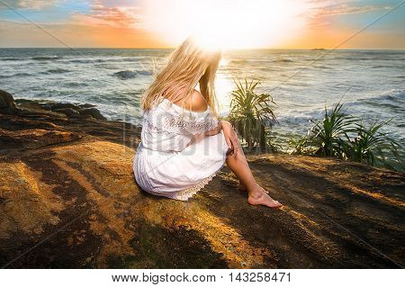 Woman sitting in the white dress on the rock in the rays of setting sun with her head in flare. Wide angle shot photo with double light exposition.