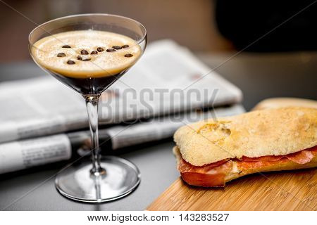 Shakerato drink in cocktail glass with panini and newspaper on the table. Italian lunch with traditional coffee and sandwich with prosciutto