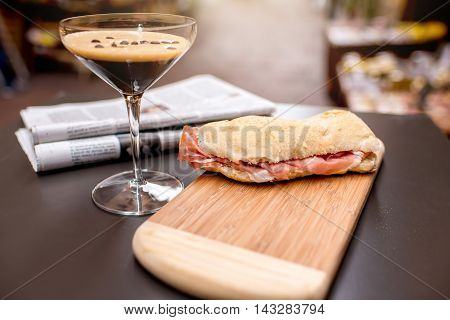 Shakerato drink in cocktail glass with panini and newspaper on the table. Italian lunch with traditional coffee and sandwich with prosciutto