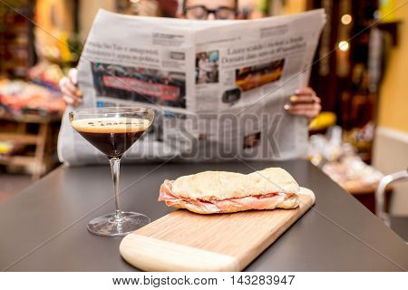 Young woman reading newspaper with shakerato drink and panini on the table outdoors on the street in italian city. Traditional italian lunch. Image with small depth of field, focused on the food