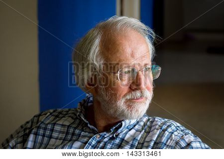 A senior man with white hair mustache beard and glasses looks contemplative as he stares out a window. His face is lit by the window light.