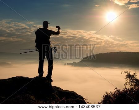Dreamy fogy landscape. Amateur photographer takes impressive photos with phone. Hiker with backpack on cliff photogrphy orange pink misty sunrise in a beautiful valley below