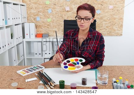 Woman painter getting ready for class in college. She is sitting in her office with cork board and mixing paint. Concept of art education and aesthetics