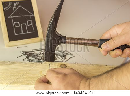 Man's hand hammering nail in wood - Image with a builder's hand hammering a small nail in a wooden board and in the background a chalkboard with a funny drawing of a house.