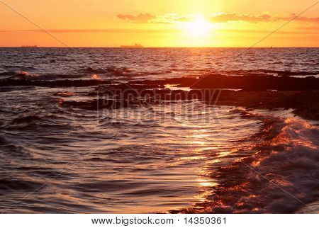 Golden sunset over en bugt, med skinnende bølger bryde i foregound og tankskibe i afstanden