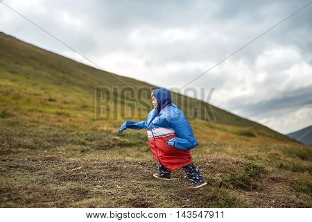 Funny kid in multi-colored big size windcheater walks on the mountain on the cloudy sky background. Shoot from the side. Horizontal.