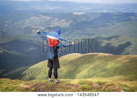 Child sits on dad's shoulders who stands on the mountain on the background of the mountains and the cloudy sky. Boy wears multi-colored big size windcheater. His father dressed in the sportswear.