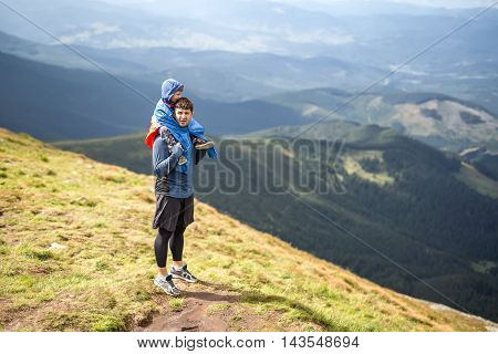 Father in sportswear stands on the mountain and holds his son on the shoulders on the background of the mountains and the cloudy sky. Boy wears multi-colored big size windcheater. Horizontal.