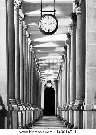 Long colonnafe corridor with columns and clock hanging from ceiling. Cloister perspective. . Black and white image.