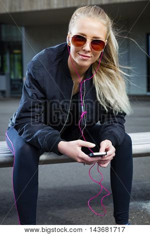 Beautiful and smiling woman with sunglasses sits on a bench in the city wearing a bomber jacket and sportswear. Listens to music and using smart phone.