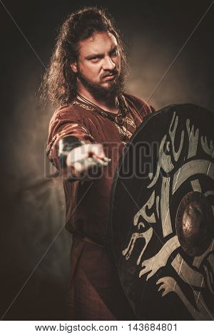 Serious viking with sword in a traditional warrior clothes, posing on a dark background.
