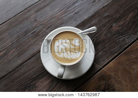 Cup of tasty latte is standing on the wooden textured table. There is nice picture is painted on the top of the latte. Latte is in the white big cup.