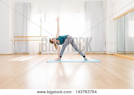 Young flexible blonde woman in utthita trikonasana triangle pose in yoga center