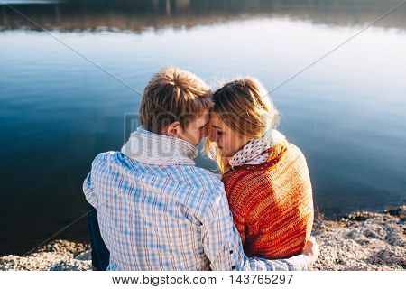 Back view of lovely young couple sitting by a lake and hugging  each other on a cold autumn day