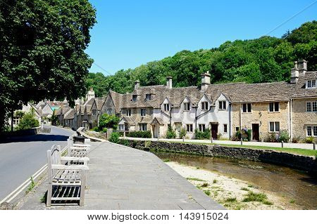 Stone cottages alongside the river Bybrook with wooden benches in the foreground Castle Combe Wiltshire England UK Western Europe.