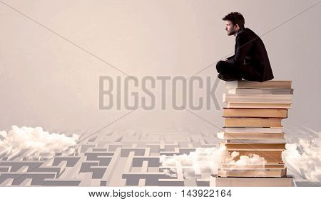 A serious businessman in suit sitting on a pile of giant books in front of a grey wall with clouds, labirynth