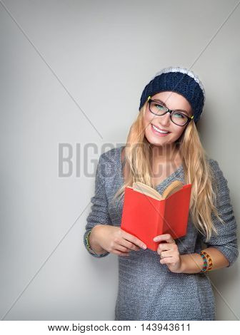 Happy student girl with book in hands wearing stylish hat and glasses isolated on gray background, enrollee of university