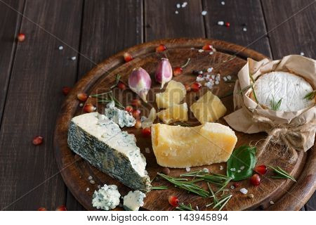 Cheese delikatessen closeup on rustic wood. Wooden round desk with blue cheese roquefort, parmesan, camembert and brie cuts decorated with garlic, pomegranate and rosemary.