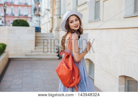 Happy beautiful young woman in hat with red backpack walking on the street