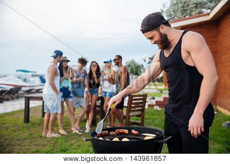 Handsome bearded young man cooking grilled sauseges and vegetables on barbeque party