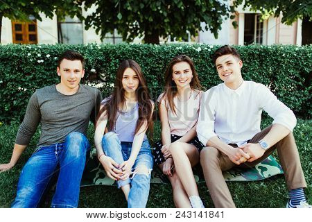 Group Of Four Young Friends In An Urban Park Sitting On The Grass In The Summer Sunshine Laughing An