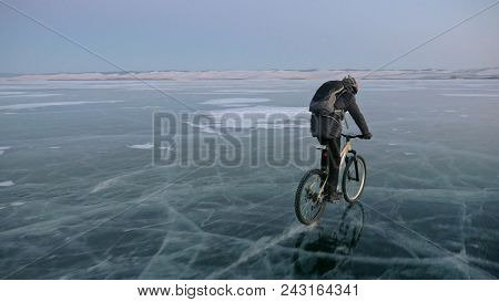 Man Is Riding A Bicycle On Ice. The Cyclist Is Dressed In A Gray Down Jacket, Backpack And Helmet. I