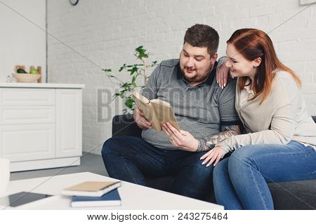 Boyfriend And Smiling Girlfriend Reading Book Together On Sofa In Living Room