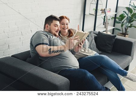 Smiling Overweight Boyfriend And Girlfriend Reading Book On Sofa At Home