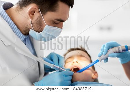 medicine, dentistry and healthcare concept - dentist and assistant with dental drill and saliva ejector treating kid patient teeth at dental clinic