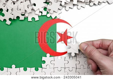 Algeria Flag  Is Depicted On A Table On Which The Human Hand Folds A Puzzle Of White Color.