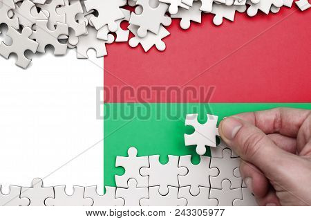 Madagascar Flag  Is Depicted On A Table On Which The Human Hand Folds A Puzzle Of White Color.
