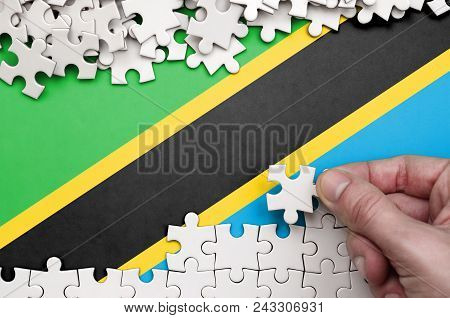 Tanzania Flag  Is Depicted On A Table On Which The Human Hand Folds A Puzzle Of White Color.