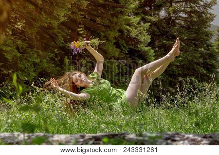 Young, Beautiful Woman In A Green Dress With Flowers In Hand Falls Into The Grass Close-up