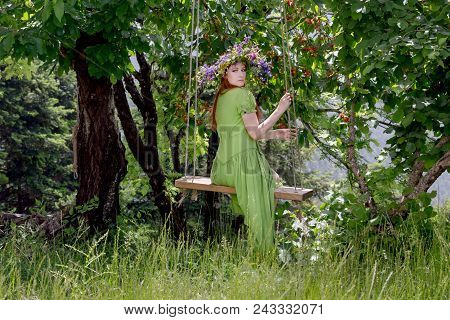 Young, Beautiful Woman In A Green Dress With A Wreath Of Wild Flowers Sits On A Homemade Swing In A 