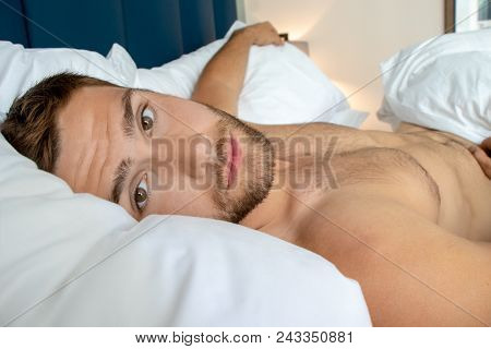 Handsome Man With Abs, Muscular, Hunky Body And Beard Lies Naked In Between White Sheets On Bed..