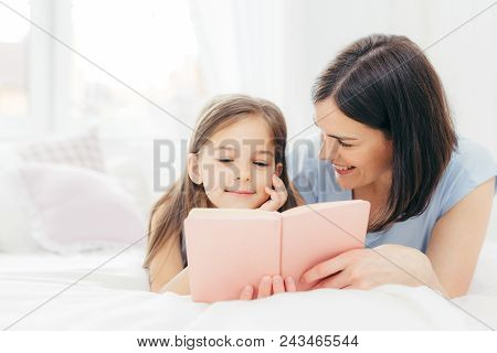 Affectionate Young Mother With Dark Hair, Looks Happily At Her Small Daughter, Read Interesting Book