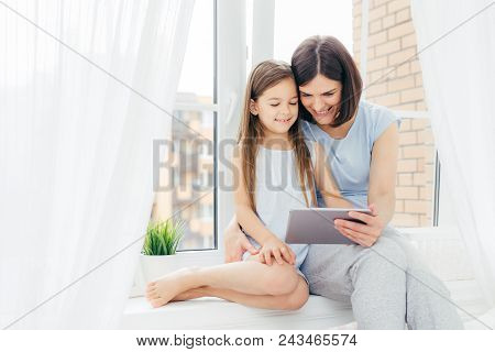 People, Technology, Family, Children Concept. Positive Young Other And Her Small Daughter Sit On Win