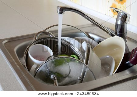 Dirty Dishes And Unwashed Kitchen Appliances Lie In Foam Water Under A Tap From A Kitchen Faucet