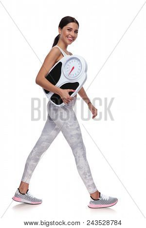 happy beautiful woman walking to side while holding scale on white background, full body picture. She is wearing white gym clothes