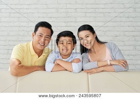 Cheerful Asian Family Leaning On Back Of Cozy Sofa And Looking At Camera With Wide Smiles While Spen