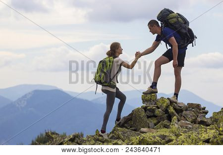 Young Tourists With Backpacks, Athletic Boy Helps Slim Girl To Clime Rocky Mountain Top Against Brig